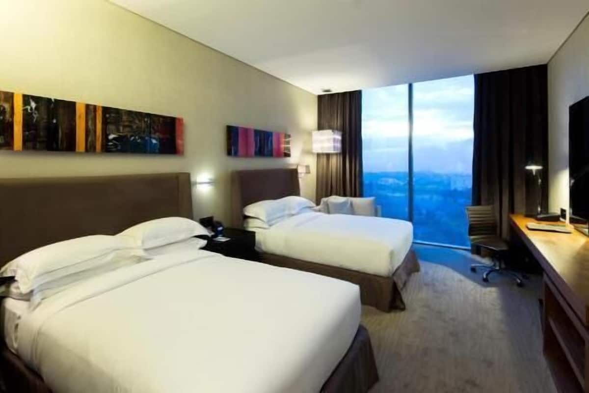 A hotel room with two beds, a desk, a chair, a TV, and a large window with a city view.