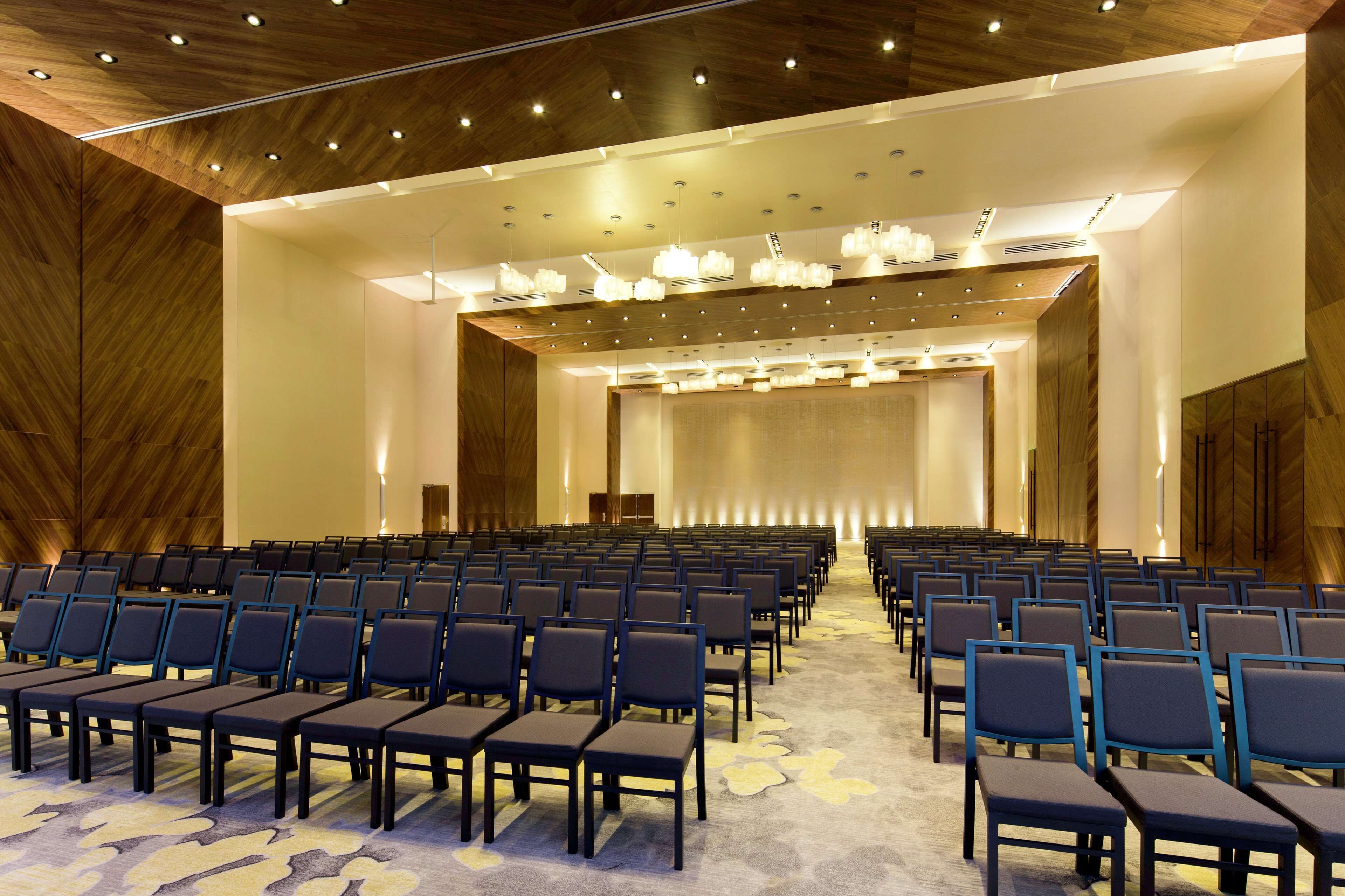 A conference room with rows of chairs, a stage, and a wooden ceiling with recessed lighting.