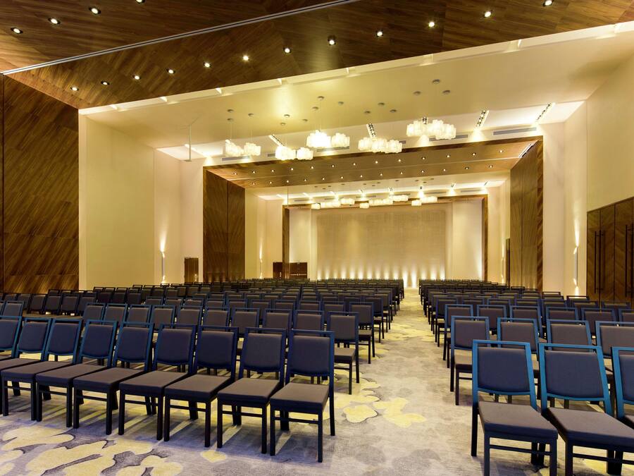 A conference room with rows of chairs, a stage, and a wooden ceiling with recessed lighting.