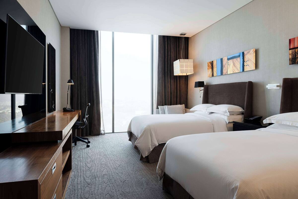 Habitación de hotel con dos camas, un escritorio con silla, televisión y un ventanal con cortinas.