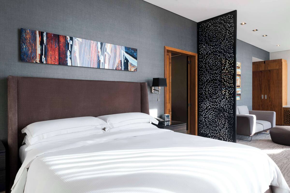 Habitación de hotel moderna con una cama grande, un escritorio con lámpara, una silla y un panel decorativo en la pared.
