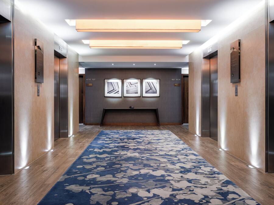 En moderne elevator foyer med et blå mønstret tæppe, elevator knapper monteret på væggen, og en loftslampe.