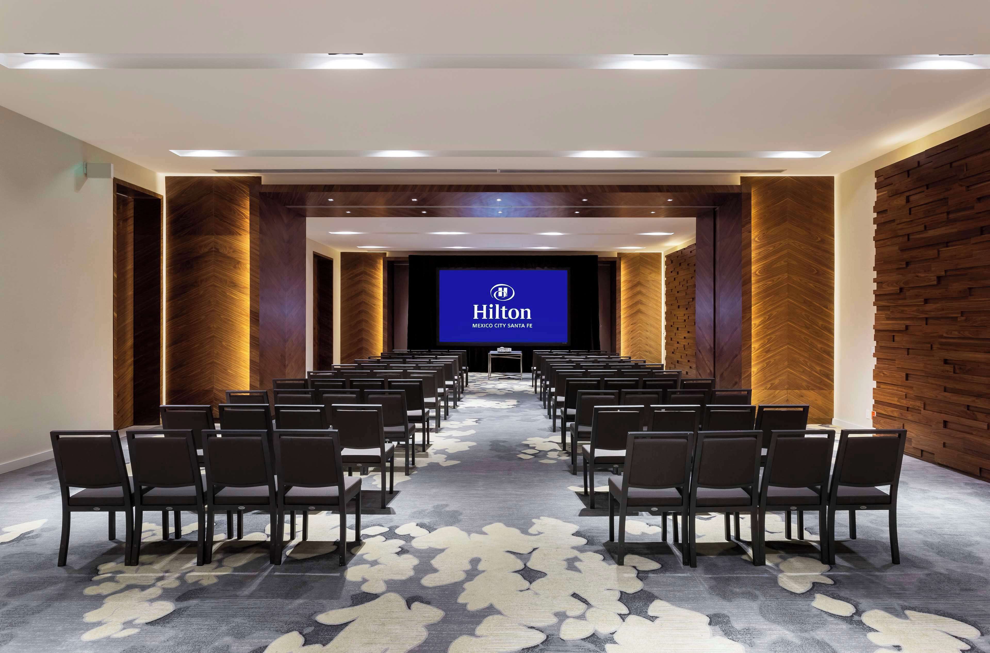 Un salón de conferencias con filas de sillas orientadas hacia una pantalla que muestra el logotipo de Hilton.
