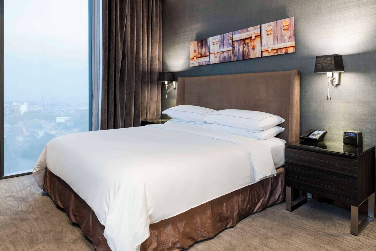 Habitación de hotel con una cama grande, una mesita de noche con teléfono y una lámpara de pared, con vistas a la ciudad.