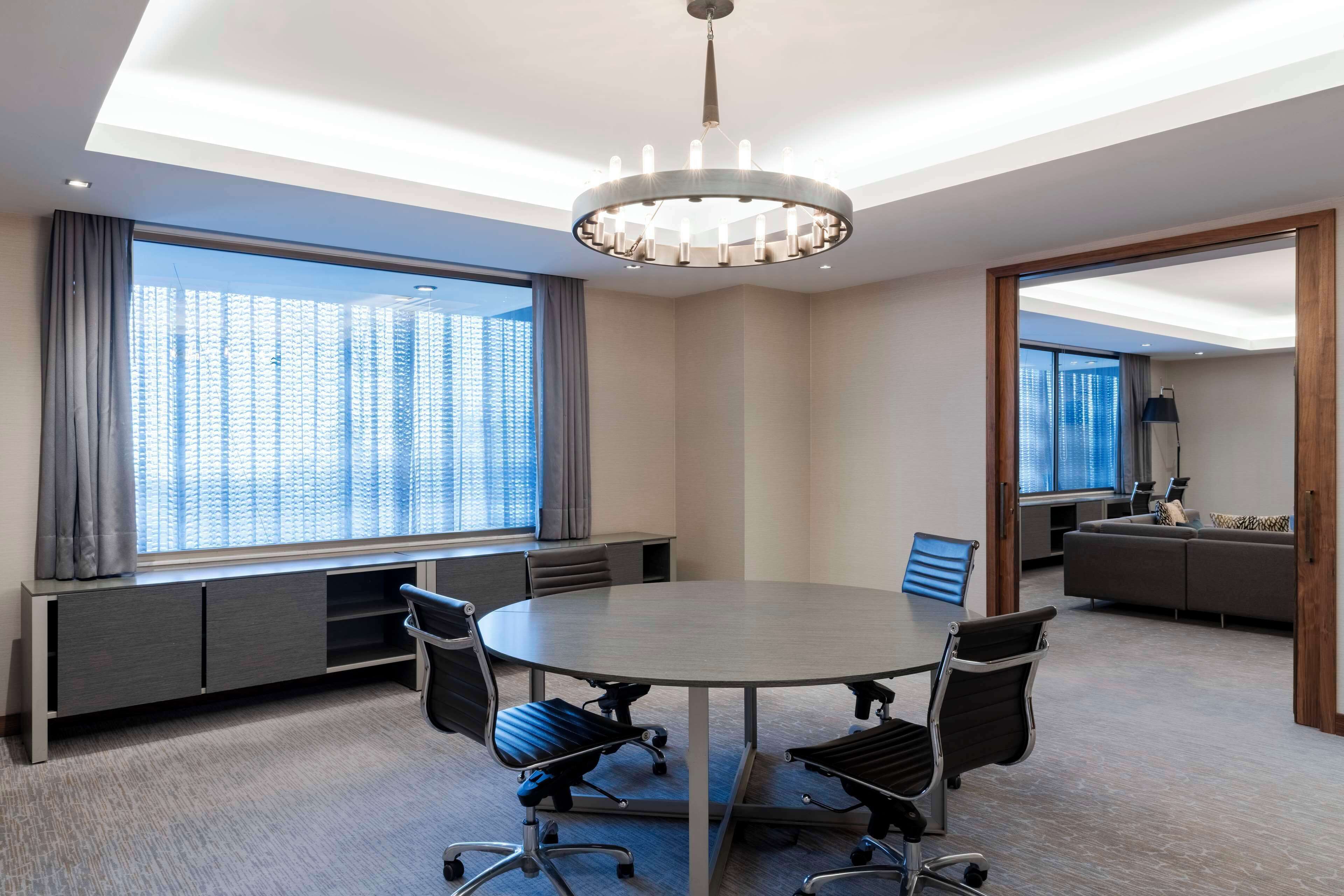 A conference room with a round table, four office chairs, a chandelier, a large window with curtains, a built-in bench, and a sofa in the adjacent room.