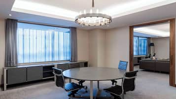 A conference room with a round table, four office chairs, a chandelier, a large window with curtains, a built-in bench, and a sofa in the adjacent room.