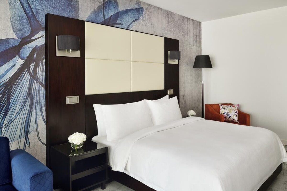 Una habitación de hotel moderna con una cama grande, una mesita de noche con un jarrón de flores, un sillón azul y una pared con un diseño abstracto en azul.