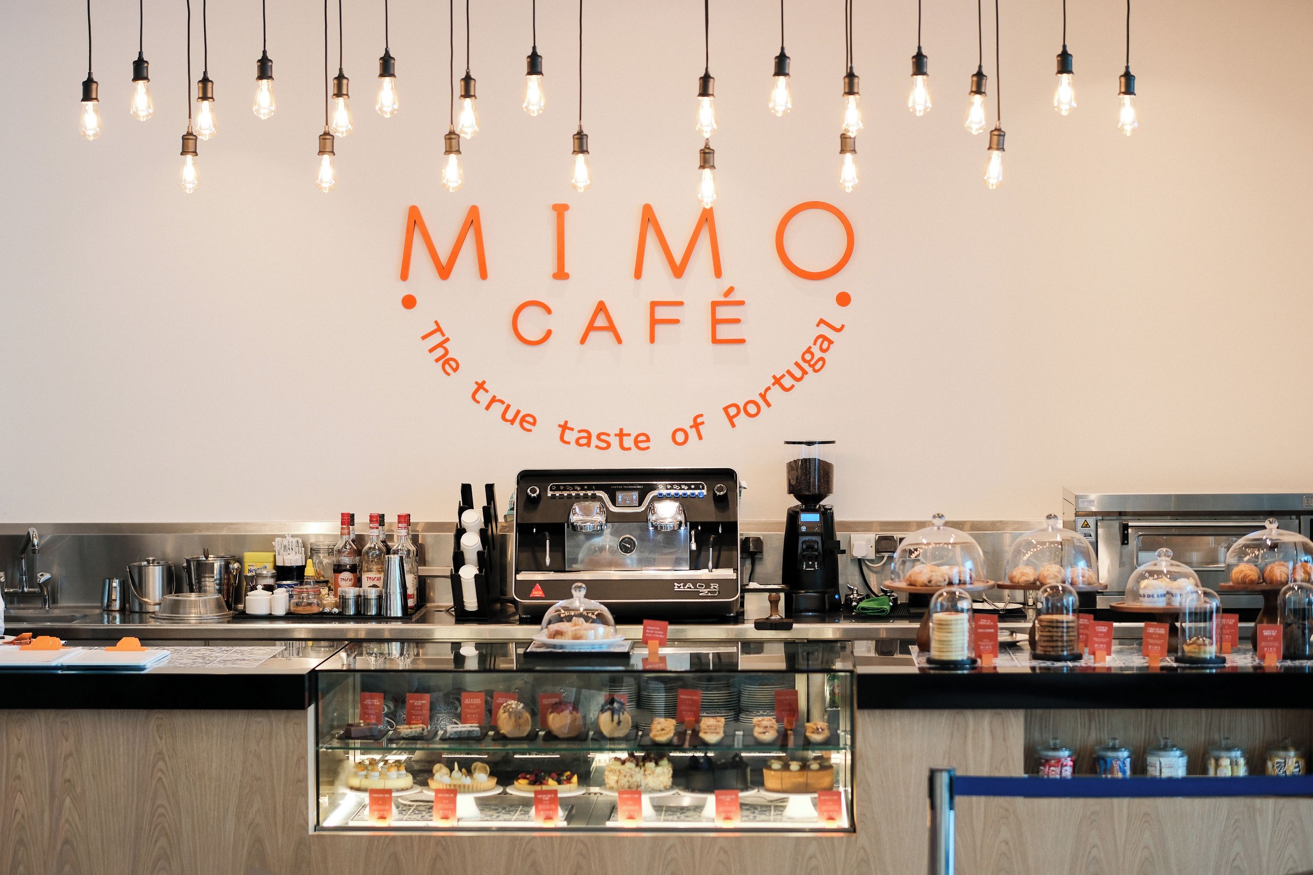A café interior with a coffee machine, display case with pastries, and hanging light bulbs.