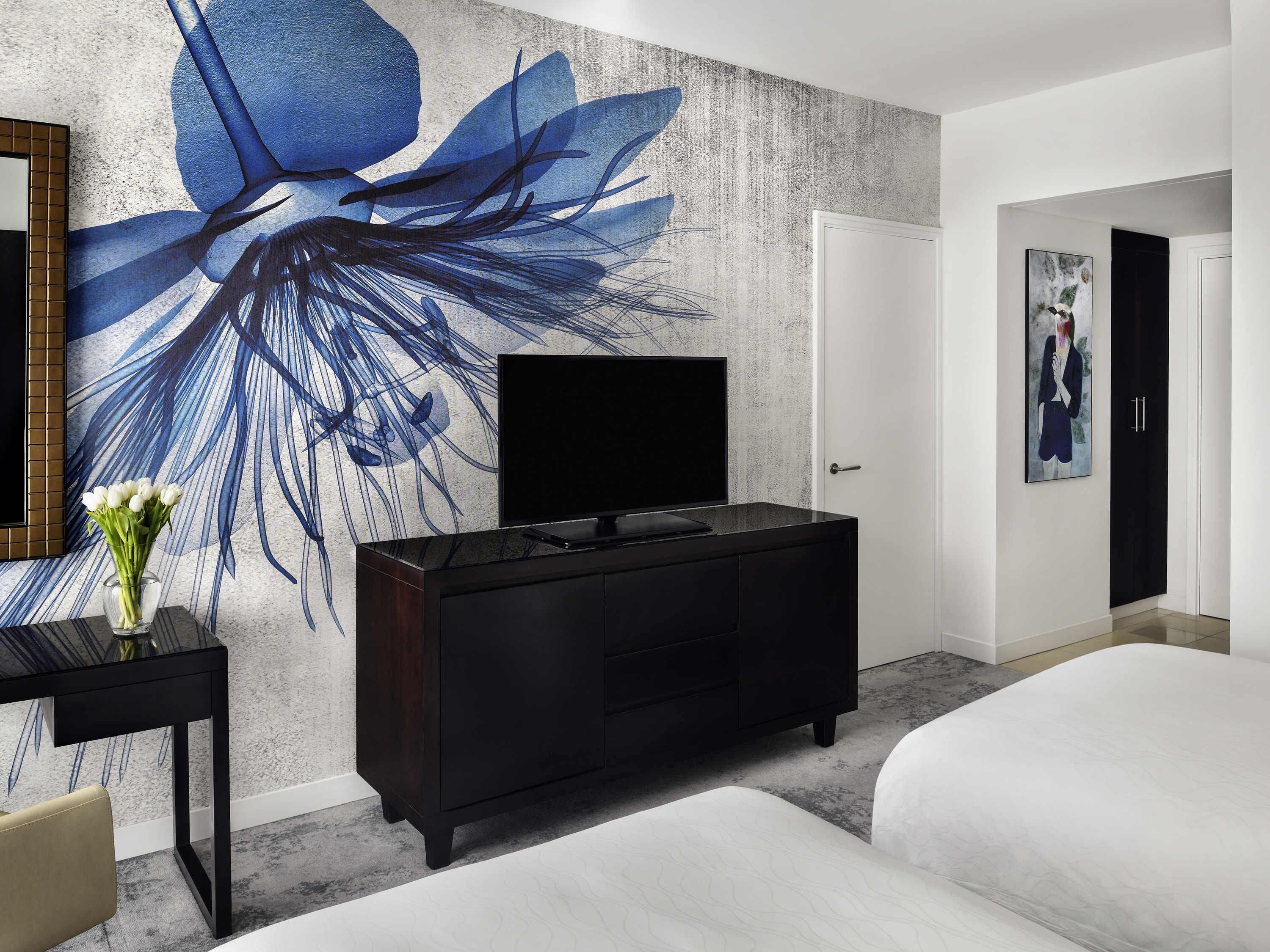 A hotel room with a large wall mural, a flat-screen TV, a black dresser, a white bed, and a small table with a vase of flowers.