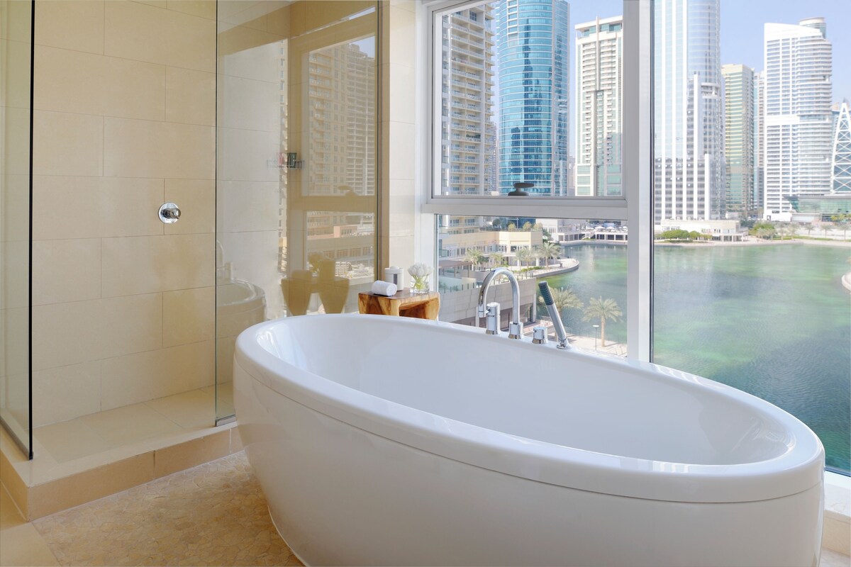 Un baño moderno con una bañera independiente, cabina de ducha con mampara de vidrio y vistas a la ciudad y al agua.