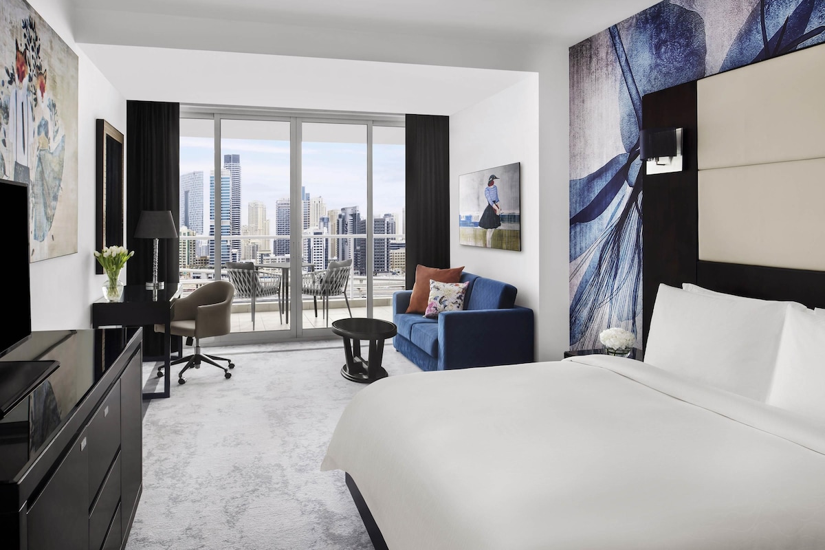 Una habitación de hotel con una cama grande, un escritorio con una silla, un sofá y vistas a la ciudad a través de amplias ventanas.
