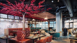 Un restaurante con una zona central de mesas donde destaca un árbol de hojas rojas, rodeado de diversas mesas y sillas para comer.
