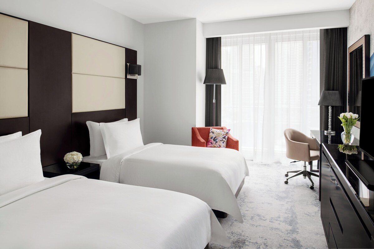 Habitación de hotel con dos camas, un escritorio, una silla y un ventanal con cortinas translúcidas.