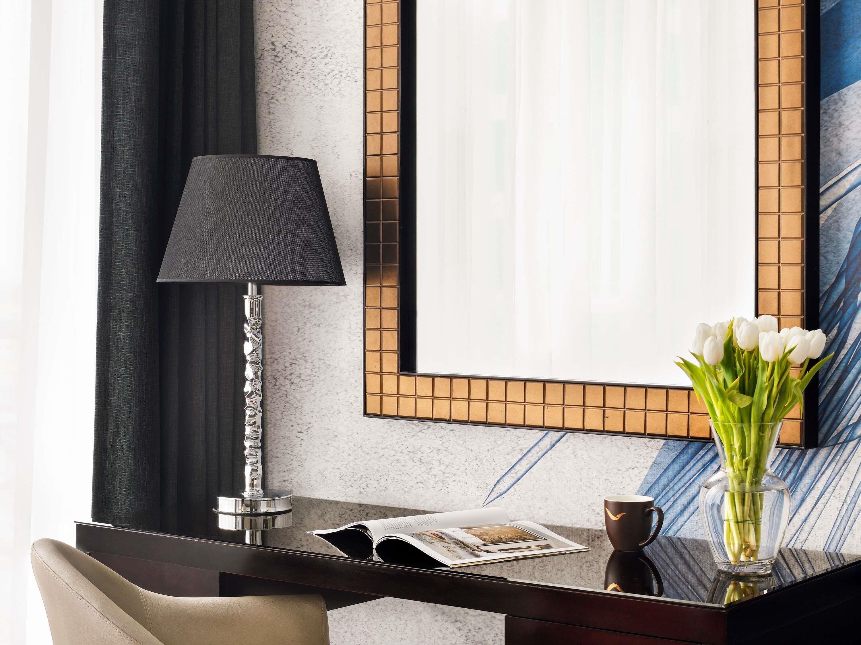 Habitación de hotel con escritorio, lámpara, un florero con flores blancas y un cuadro en la pared.