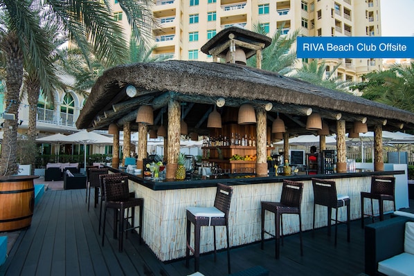 A RIVA Beach Club Offsite with a thatched roof bar area, wicker chairs, and a bar stocked with various beverages.