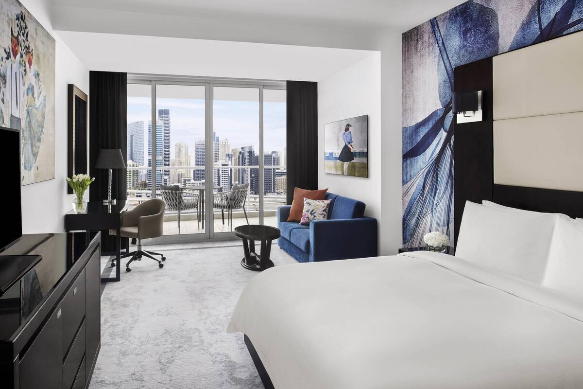 A hotel room with a large bed, a desk with a chair, a sofa, and a view of the cityscape through large windows.