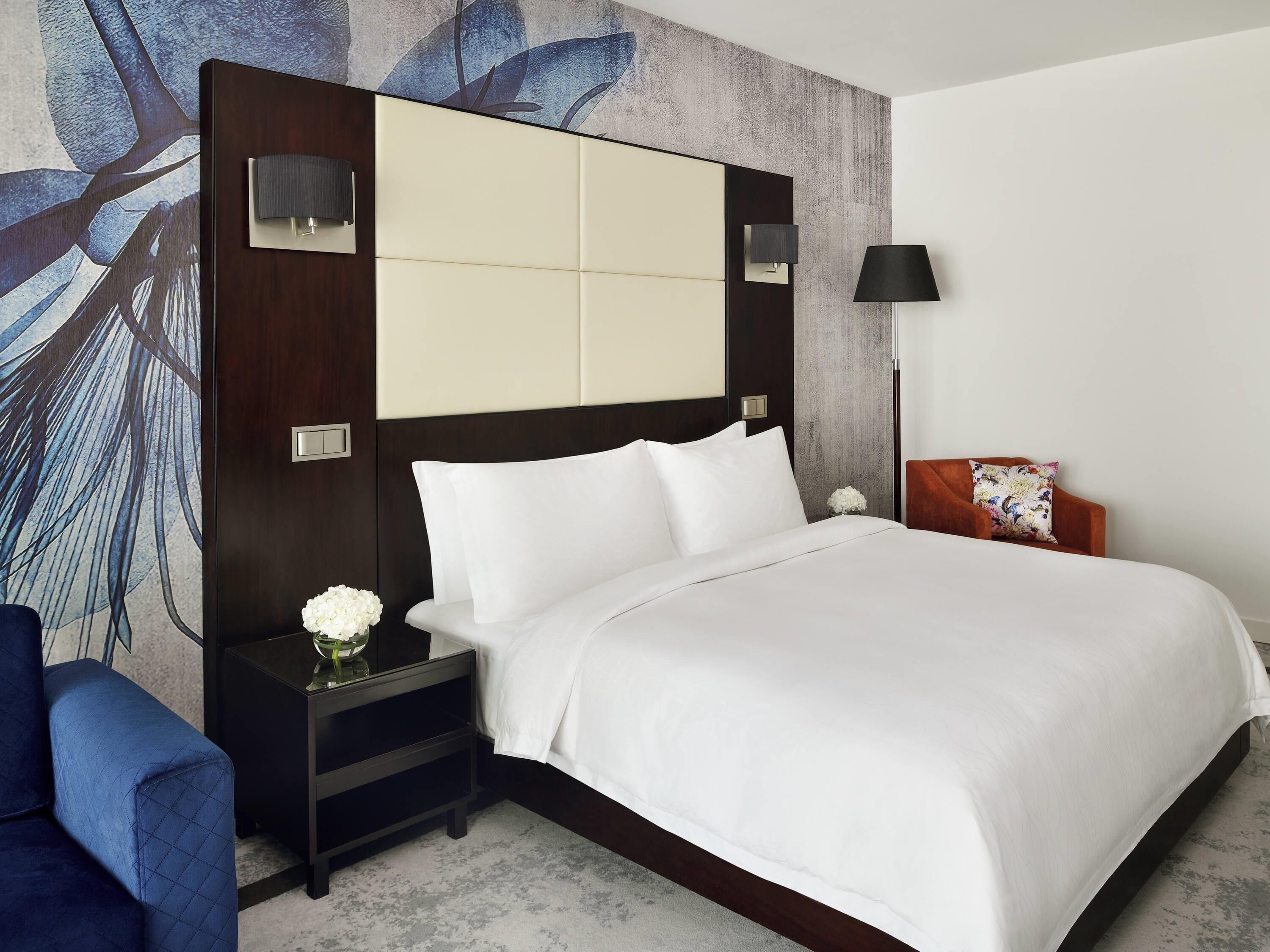 Habitación de hotel con una cama grande, una mesita de noche con un florero y un sillón azul, y una pared con un diseño de hojas.