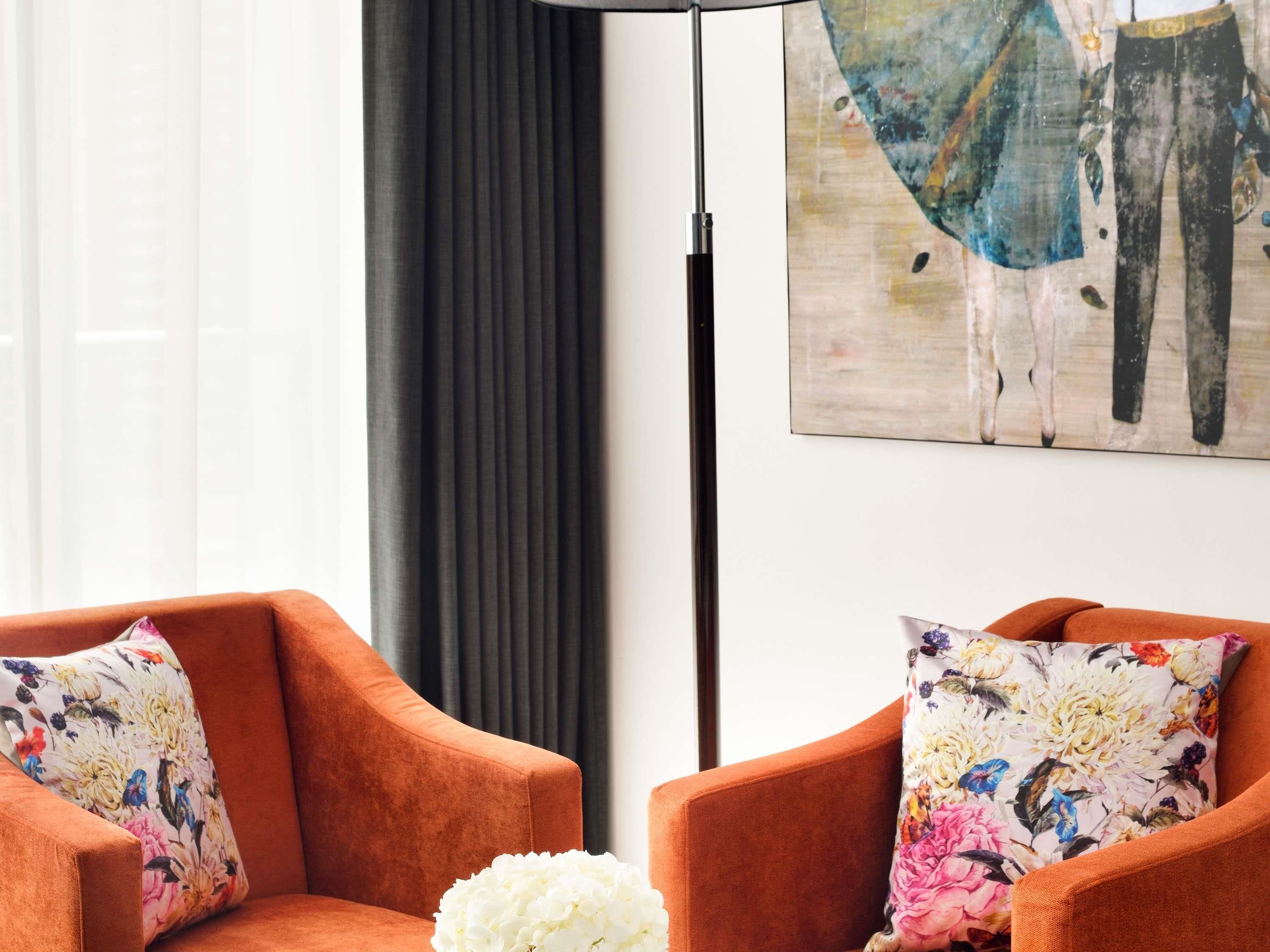 A room with two orange armchairs, a white vase, and a painting on the wall.