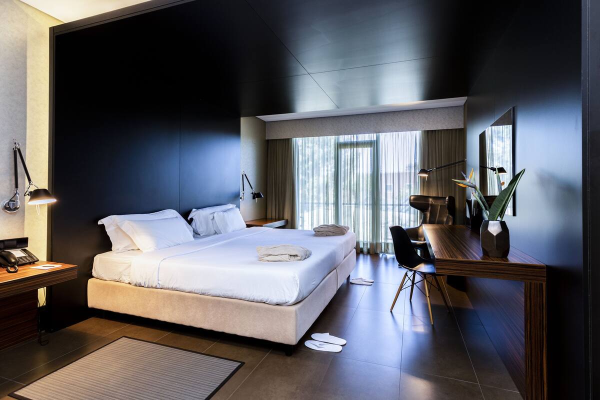 Een moderne hotelkamer met een groot bed, bedlampen, een bureau met telefoon en uitzicht op het omliggende landschap door grote ramen.