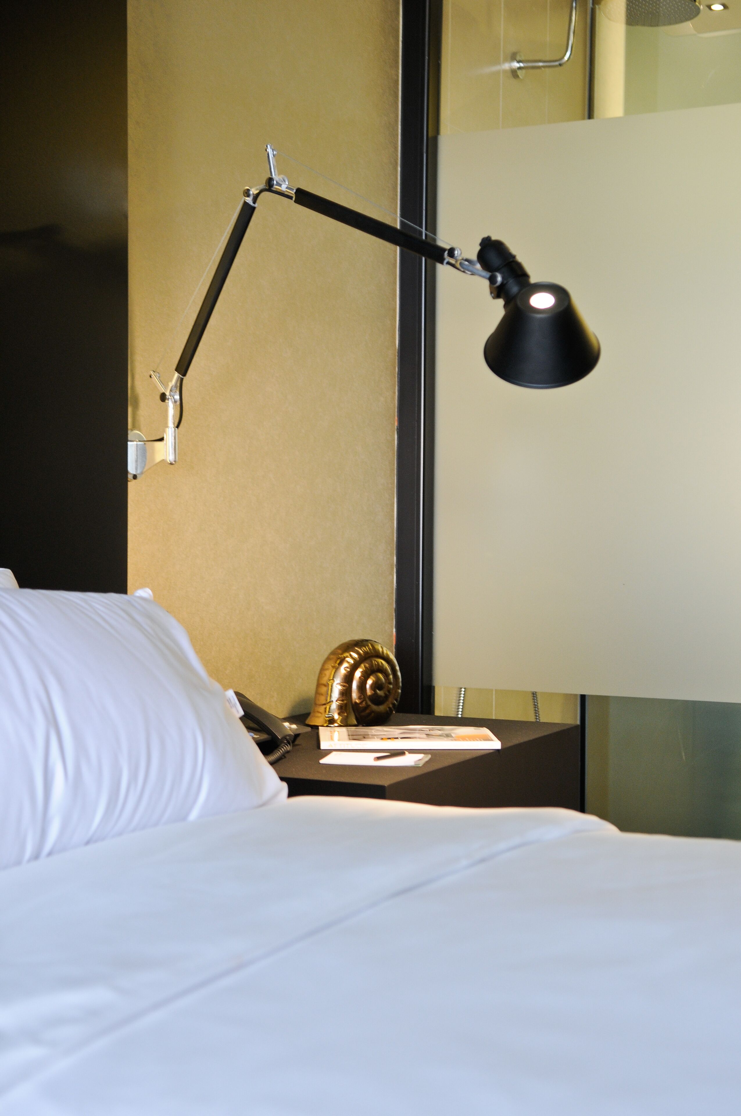Un lit avec du linge de lit blanc, une lampe de bureau noire, une petite figurine d’escargot et un miroir au mur.