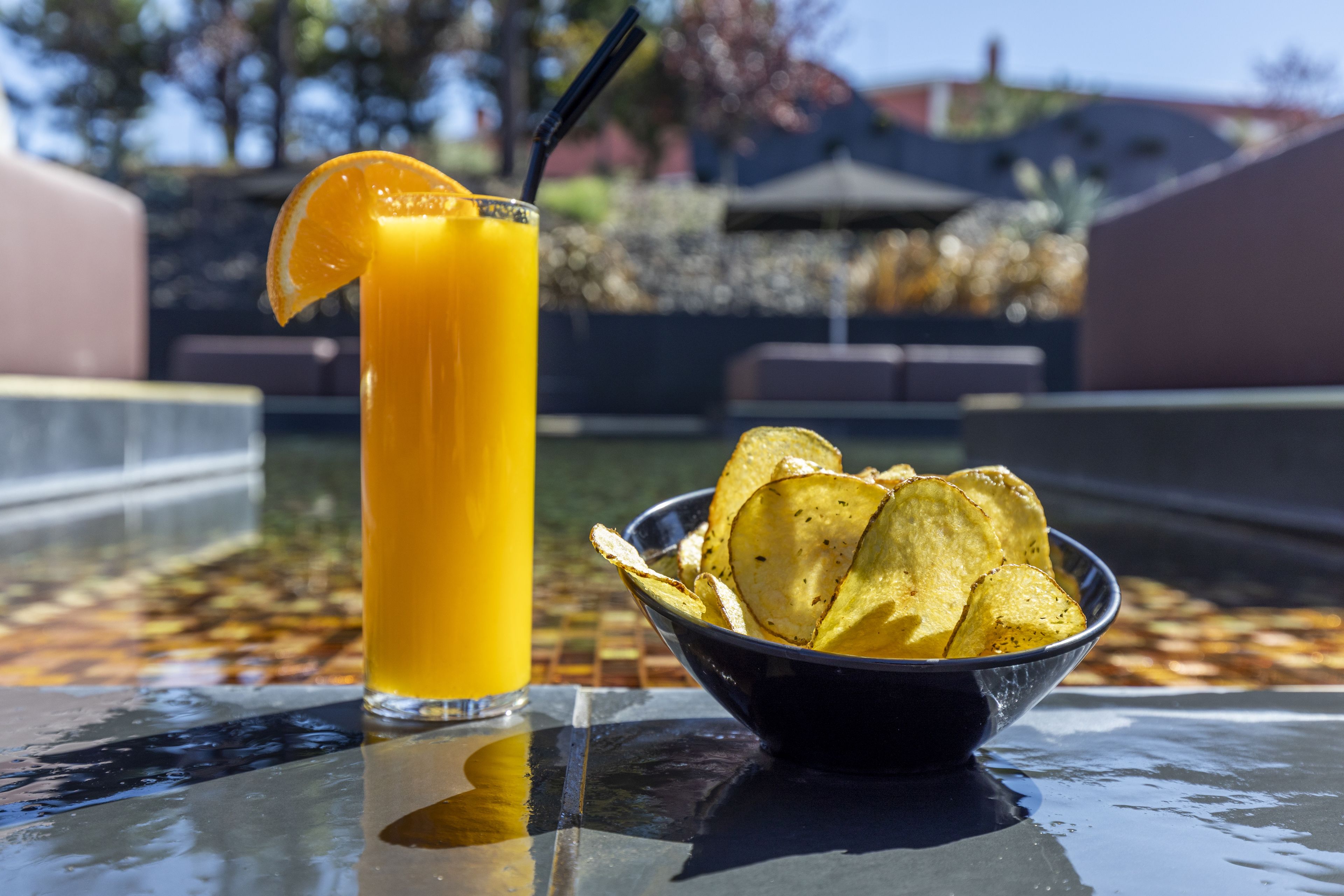 Un verre de jus d’orange, garni d’une tranche de citron et agrémenté d’une paille noire, à côté d’un bol de chips sur une surface réfléchissante.