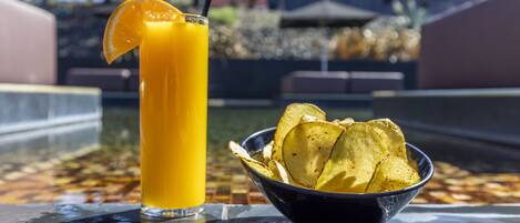 Un verre de jus d’orange, garni d’une tranche de citron et agrémenté d’une paille noire, à côté d’un bol de chips sur une surface réfléchissante.