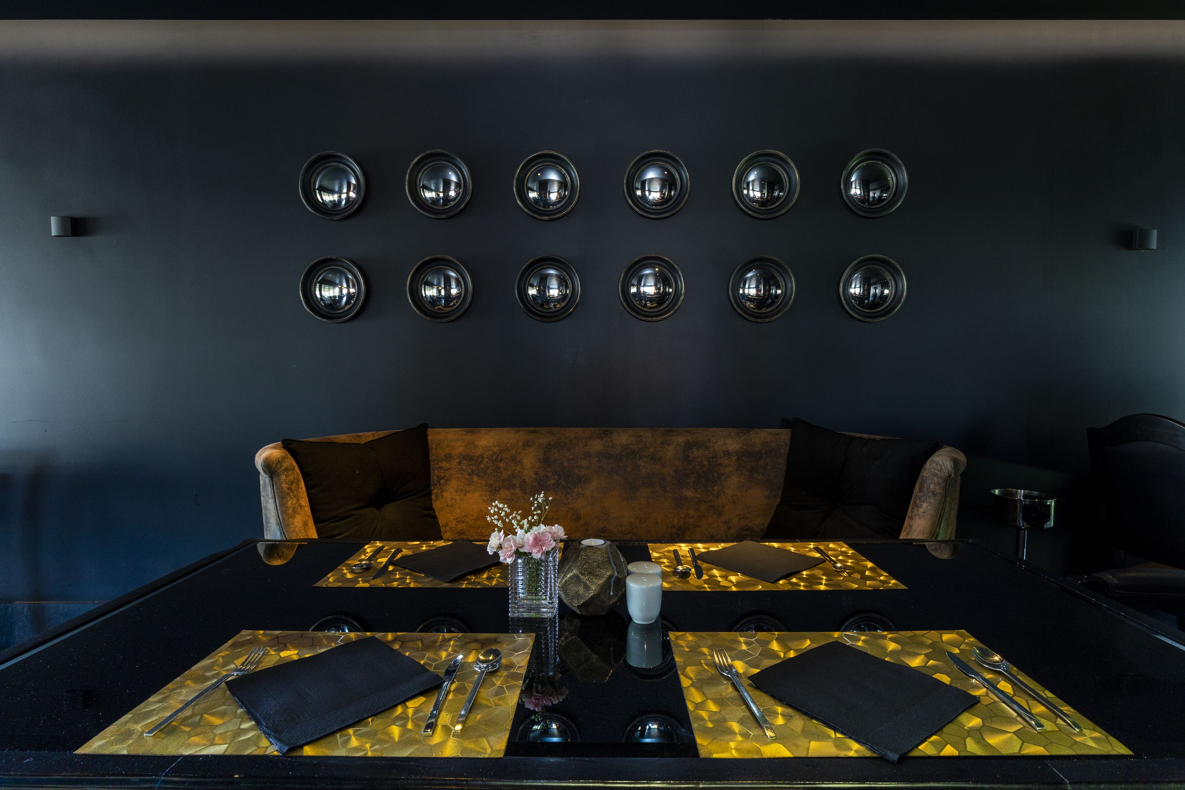 Un comedor moderno con una mesa negra para dos, un florero con flores y una pared con espejos circulares.