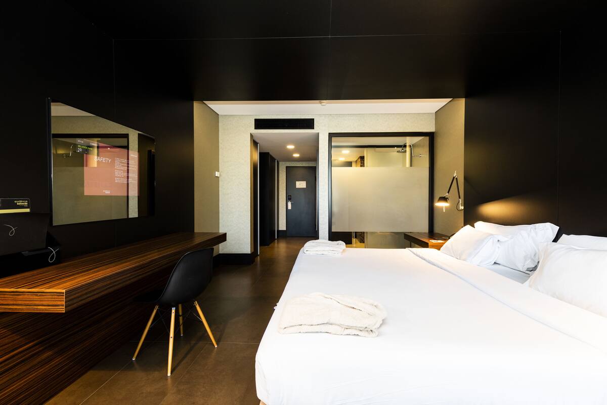 Een hotelkamer met een houten bureau, een bed met witte lakens, een stoel en een nachtkastje.