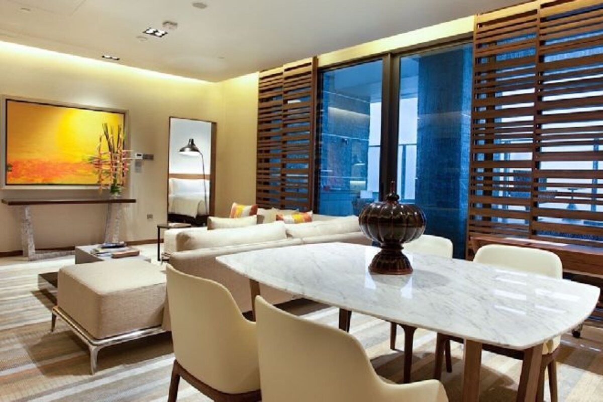 Una habitación de hotel moderna con una mesa de comedor de mármol, un conjunto de sofás, un cuadro grande y vistas a través de una ventana con persianas de madera.
