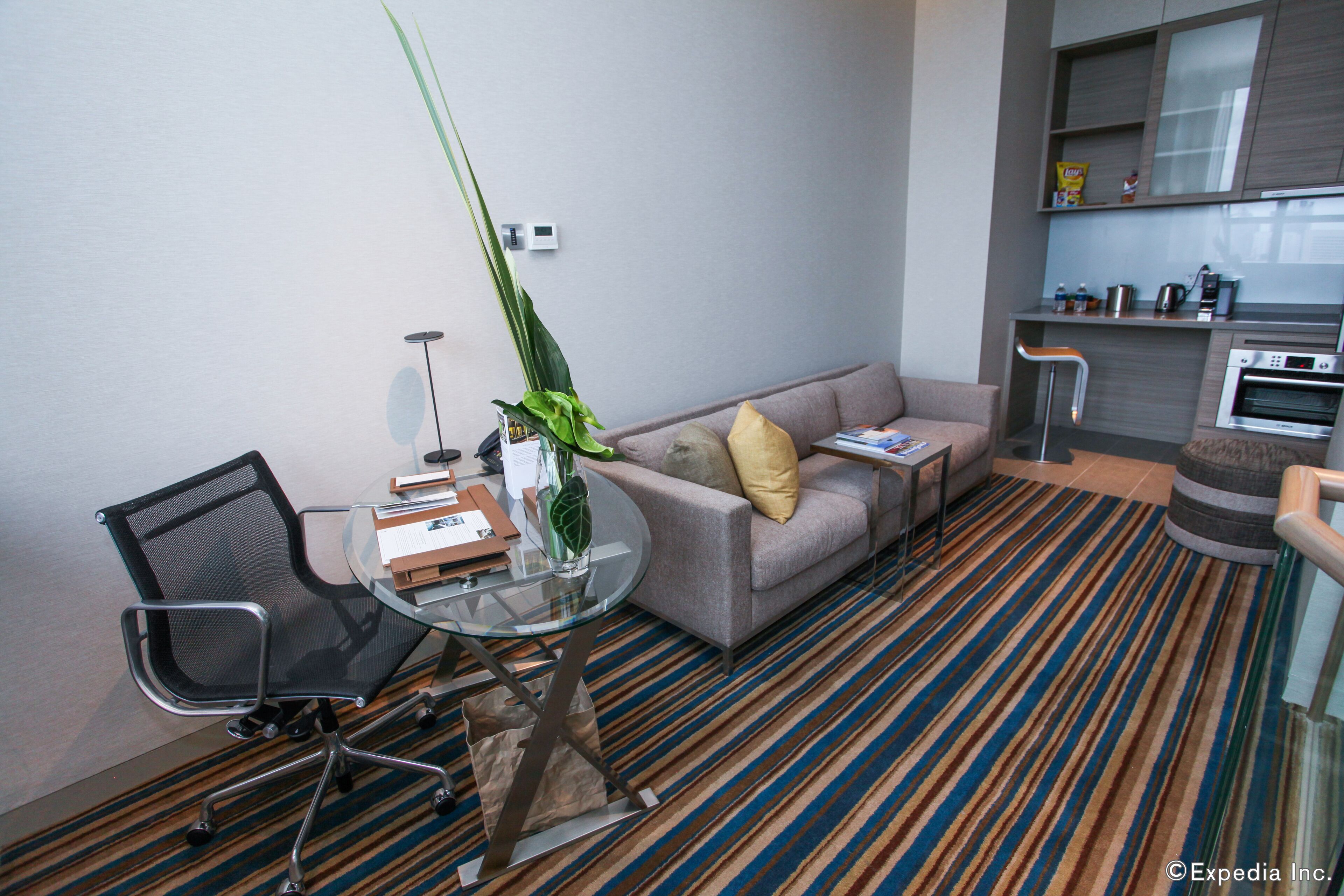 Ein modernes Wohnzimmer mit einer Couch, einem Glastisch, einem schwarzen Bürostuhl, einem Streifenmuster-Teppich und einer Küchenzeile mit Mikrowelle und Kaffeemaschine.