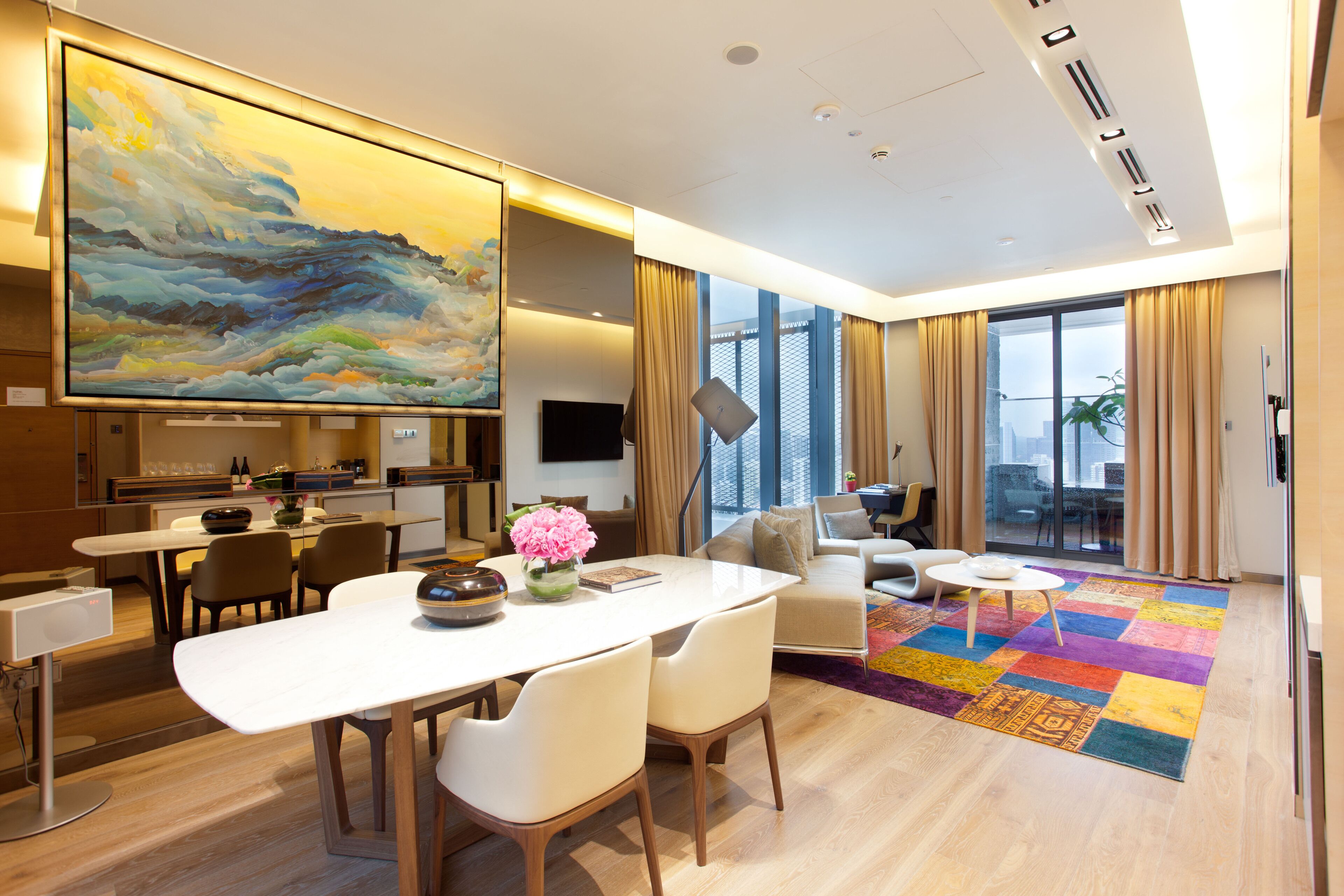 Habitación de hotel moderna con zona de comedor, un cuadro grande, un televisor de pantalla plana y vistas de la ciudad a través de amplias ventanas.