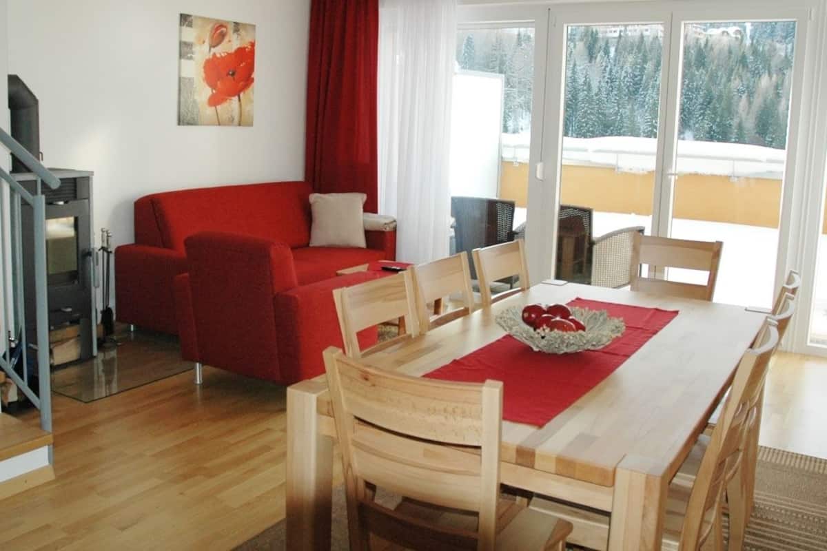 A modern living room with a red sofa, wooden dining table, and chairs. There is a painting on the wall, a window with a view of snow-covered trees, and a balcony with outdoor furniture.