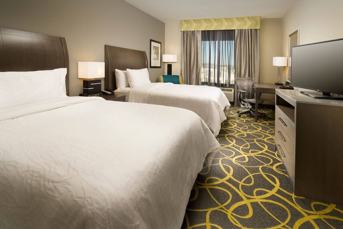 A hotel room with two beds, a desk, a chair, a television, and a window with curtains.