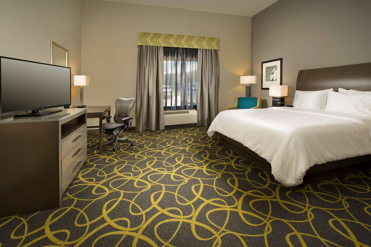 A hotel room with a large bed, a desk with a flat-screen TV, a chair, a desk lamp, and a window with curtains.