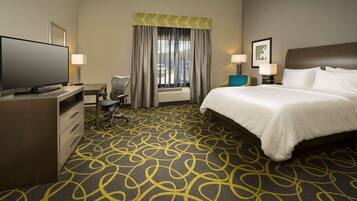A hotel room with a large bed, a desk with a flat-screen TV, a chair, a desk lamp, and a window with curtains.