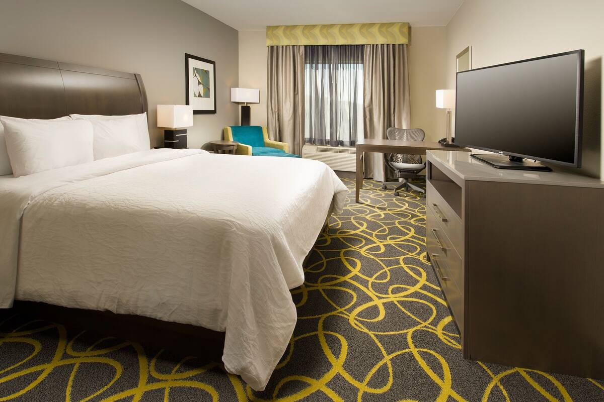 A hotel room with a large bed, a desk with a flat-screen TV, a chair, and a desk lamp.