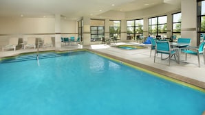 An indoor swimming pool with lounge chairs and tables, large windows, and a tiled floor.