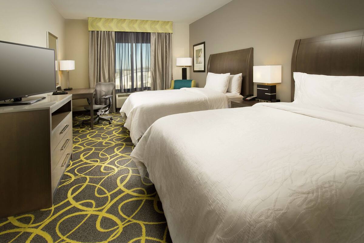 A hotel room with two beds, a desk with a television, a chair, and a window with curtains.