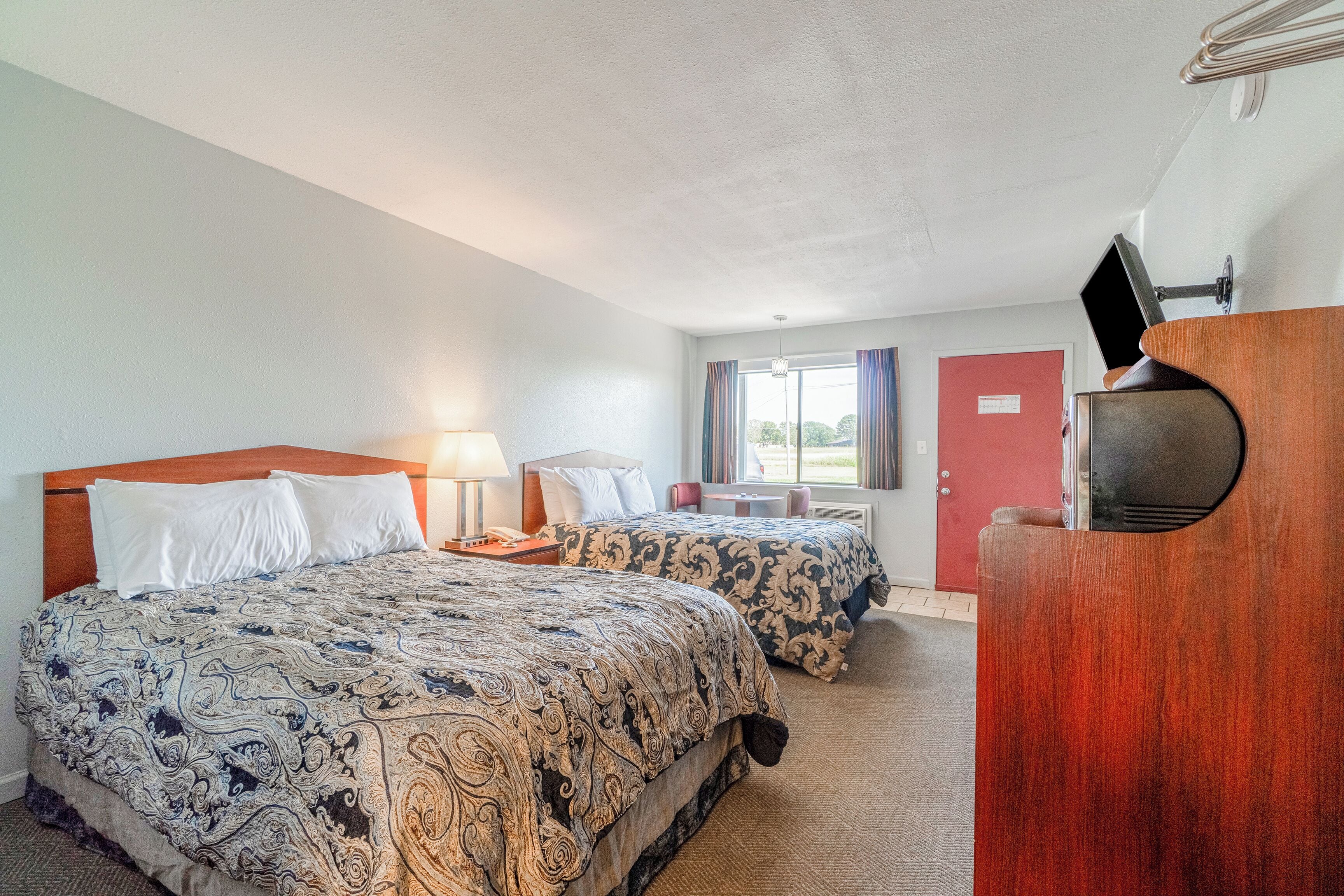 A hotel room with two beds, a desk, a chair, a lamp, and a TV.