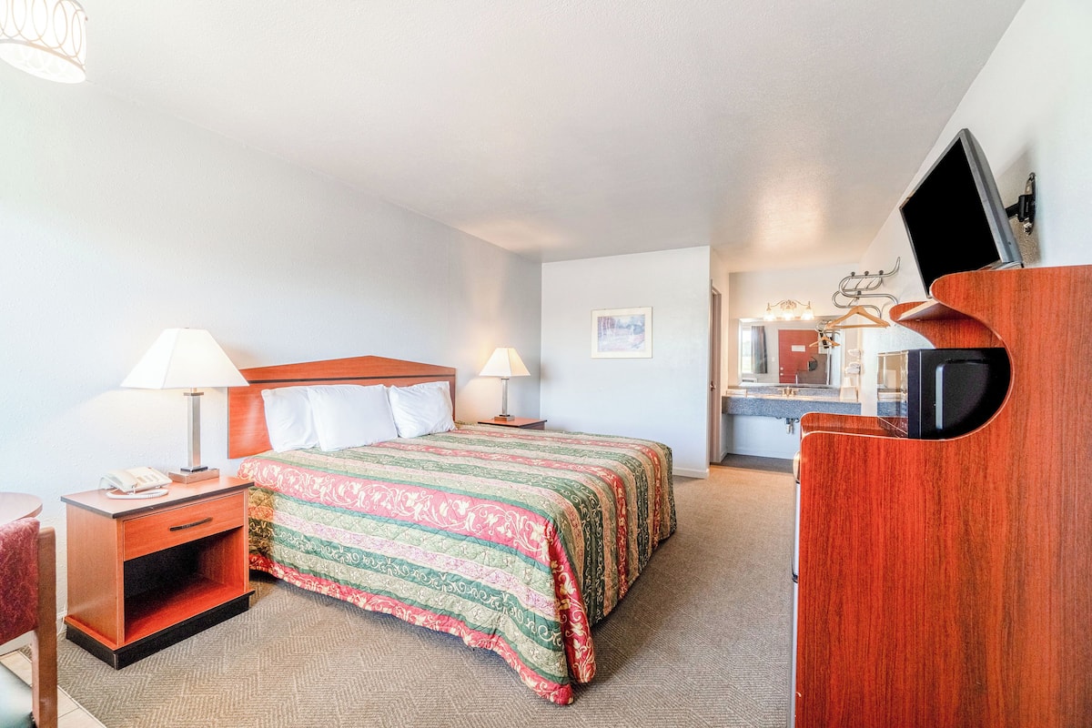 Quarto de hotel com uma cama, duas mesas de cabeceira com luminárias, uma cômoda de madeira, uma televisão e um banheiro visível ao fundo.