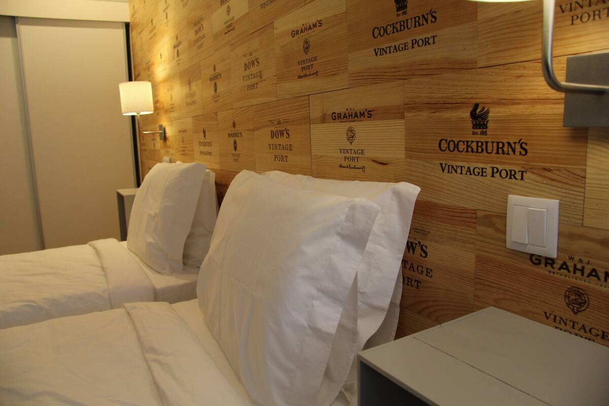 A hotel room with two beds, a wooden wall with vintage port names, a nightstand, and a lamp.