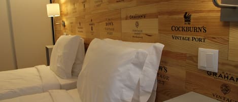 A hotel room with two beds, a wooden wall with vintage port names, a nightstand, and a lamp.