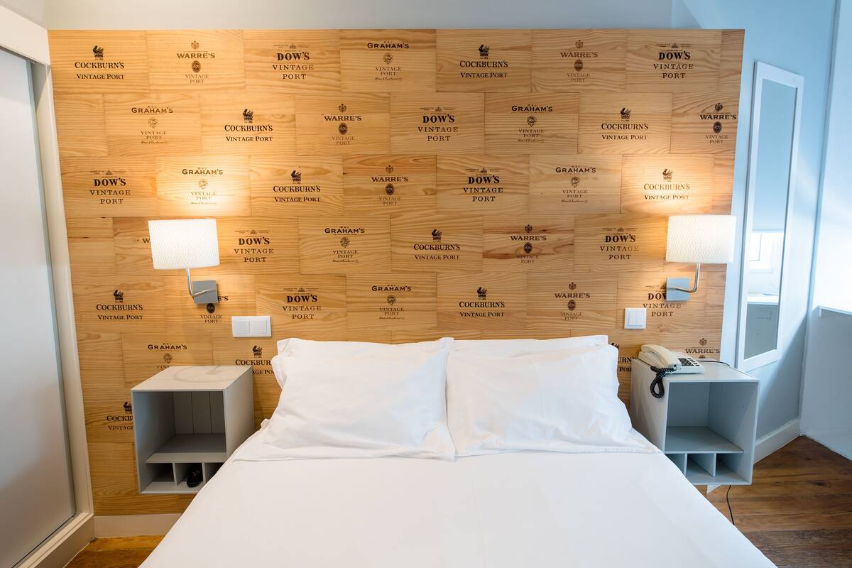A hotel room with a wooden headboard displaying brand names, a bed with white linens, bedside tables with lamps, and a small shelf.