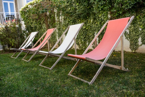 A row of lounge chairs with metal frames and fabric seats, arranged on a grassy lawn with a building and greenery in the background.