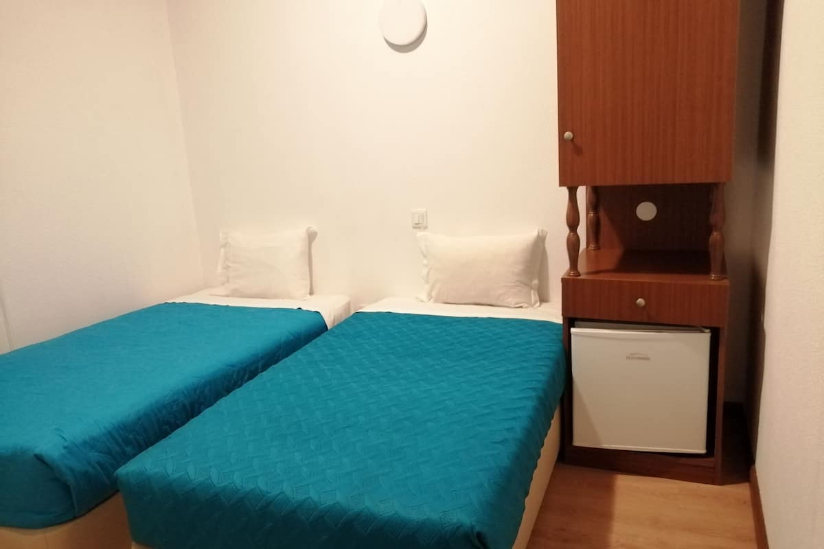 A small, clean room with two single beds, a wooden cabinet, and a small refrigerator.