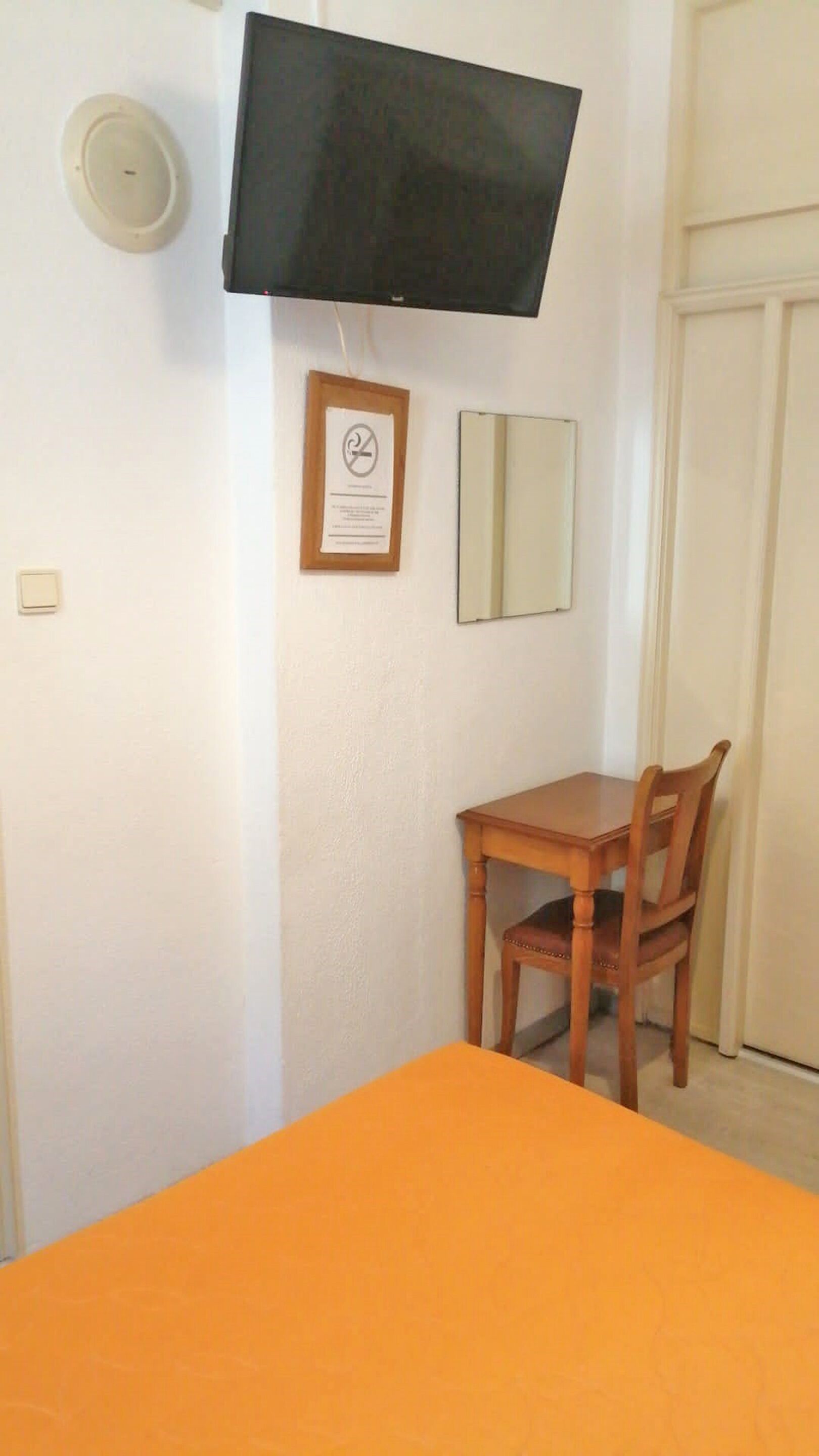 Een kamer met een aan de muur bevestigde televisie, een ingelijst certificaat, een spiegel en een houten stoel bij een klein tafeltje.