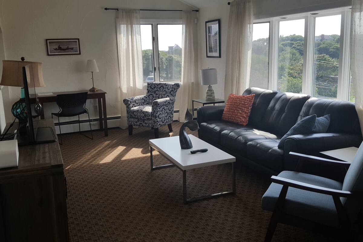 Un salon avec un canapé en cuir noir, un fauteuil à motifs et une table basse blanche. On y trouve un bureau avec une lampe, un tableau encadré au mur et de grandes fenêtres avec des rideaux.