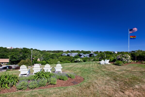 A grassy area with white Adirondack chairs, a flagpole with the American flag, and a row of houses in the background.