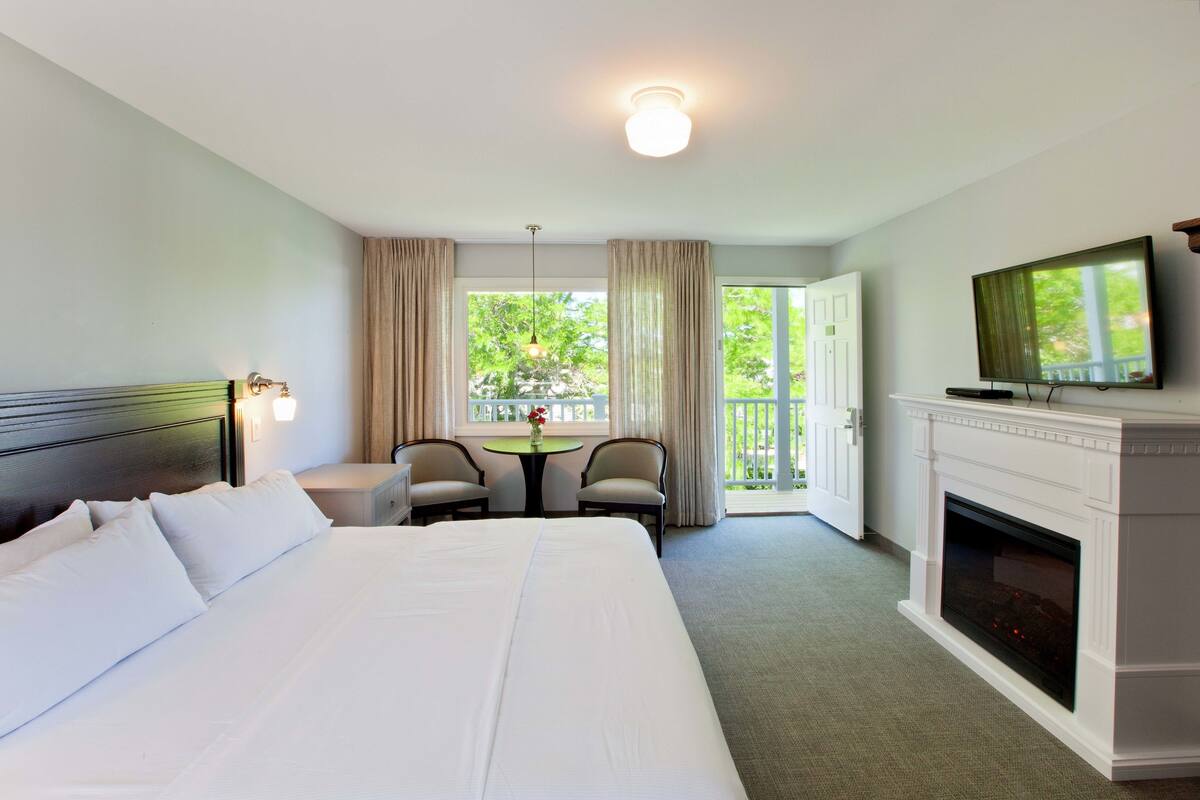 Une chambre d’hôtel avec un grand lit, un téléviseur fixé au-dessus d’une cheminée, une petite table avec des chaises et un balcon avec des rideaux.