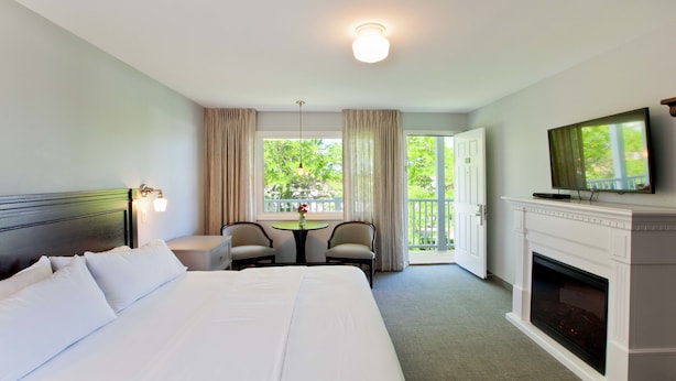 A hotel room with a large bed, a television mounted above a fireplace, a small table with chairs, and a balcony with curtains.